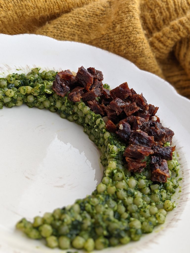 A plate of soft and bite sized kale pesto pasta arranged in a crescent shape and topped with minced barbeque pork.